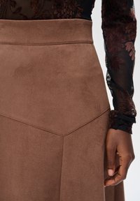 Zbliżenie na brązową zamszową spódnicę z detalami w talii i szwie, noszoną z czarną, przeźroczystą, kwiecistą bluzką z długim rękawem oraz widoczną prawą ręką na boku.