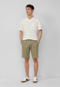 Leichtes, strukturiertes weißes Polo-Shirt mit Kragen und Tasche, kombiniert mit khakifarbenen Shorts. Sneaker mit braunen Akzenten runden das Outfit ab.