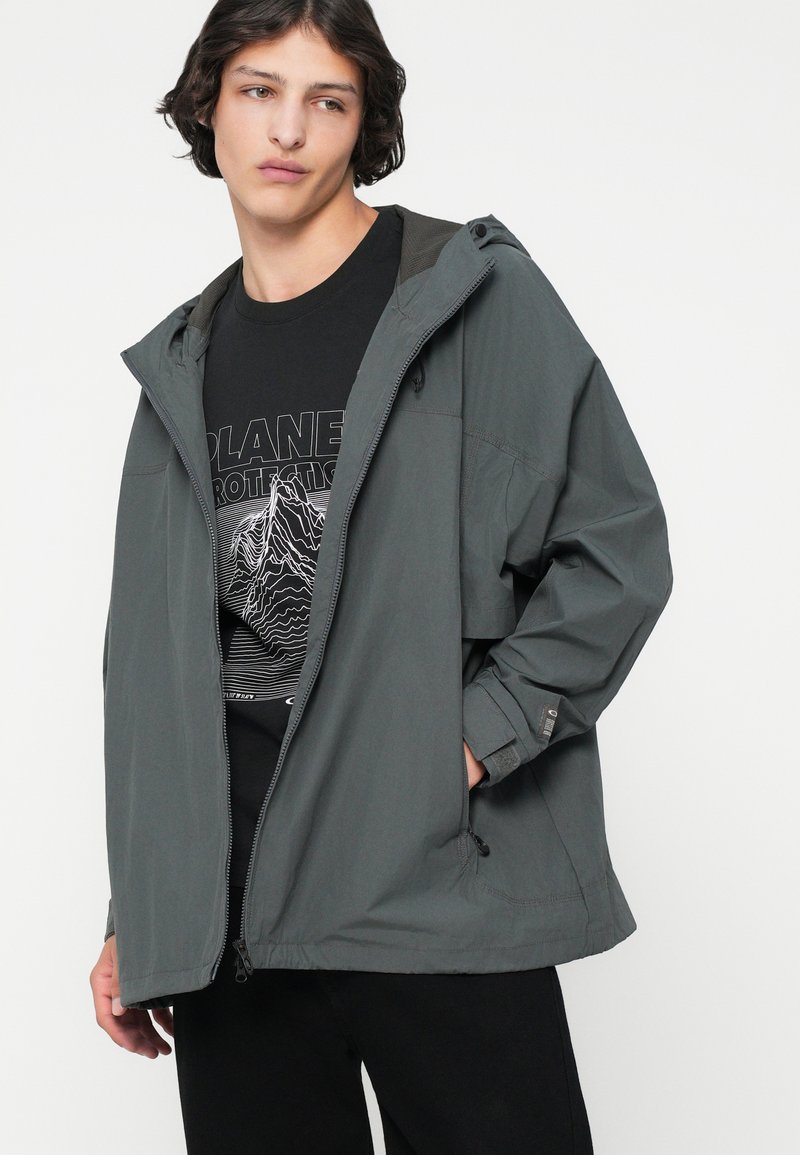 Jong persoon in een grijze hoodie over een zwart t-shirt met de tekst "Planet Protection", staand met één hand in de zak tegen een witte achtergrond.