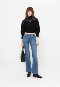 Pull court noir avec détails en dentelle, associé à un jean flare bleu clair et des bottines noires. Tient un sac tote noir.