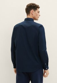 Marineblauwe langemouwen blouse met een knoopsluiting kraag, met een soepele textuur en een eenvoudig ontwerp. Achteraan is de gebogen zoom en de geknoopte manchetten zichtbaar.