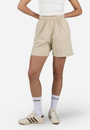 Personne portant un short beige à taille élastique, des chaussettes blanches avec texte noir, des baskets blanches Adidas avec des bandes marron, et un top court blanc.