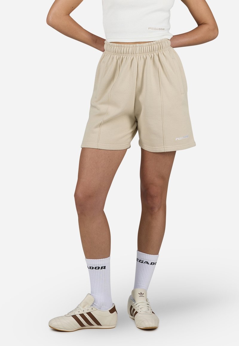 Personne portant un short beige à taille élastique, des chaussettes blanches avec texte noir, des baskets blanches Adidas avec des bandes marron, et un top court blanc.