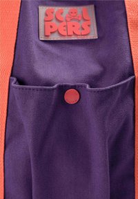 Tissu violet avec poche boutonnée rouge et patch logo rectangulaire indiquant "Scalpers" avec un crâne et des os croisés au-dessus. Bords orange.