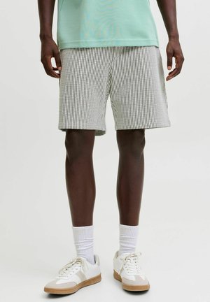 Muž oblečený v svetlozelenom tričku, sivých šortkách s textúrou, bielych ponožkách po lýtka a bielych teniskách s béžovými akcentmi.