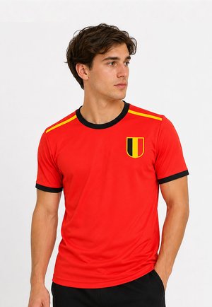 Jonge man draagt een rood sportshirt met zwarte bies, gele strepen op de schouders en een schildembleem in zwart en geel op de borst.