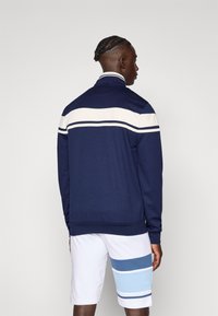 Marineblauwe sweatshirt met een ritssluiting met witte en blauwe horizontale strepen langs de schouders; geribbelde manchetten en zoom; gecombineerd met witte shorts met blauwe strepen.