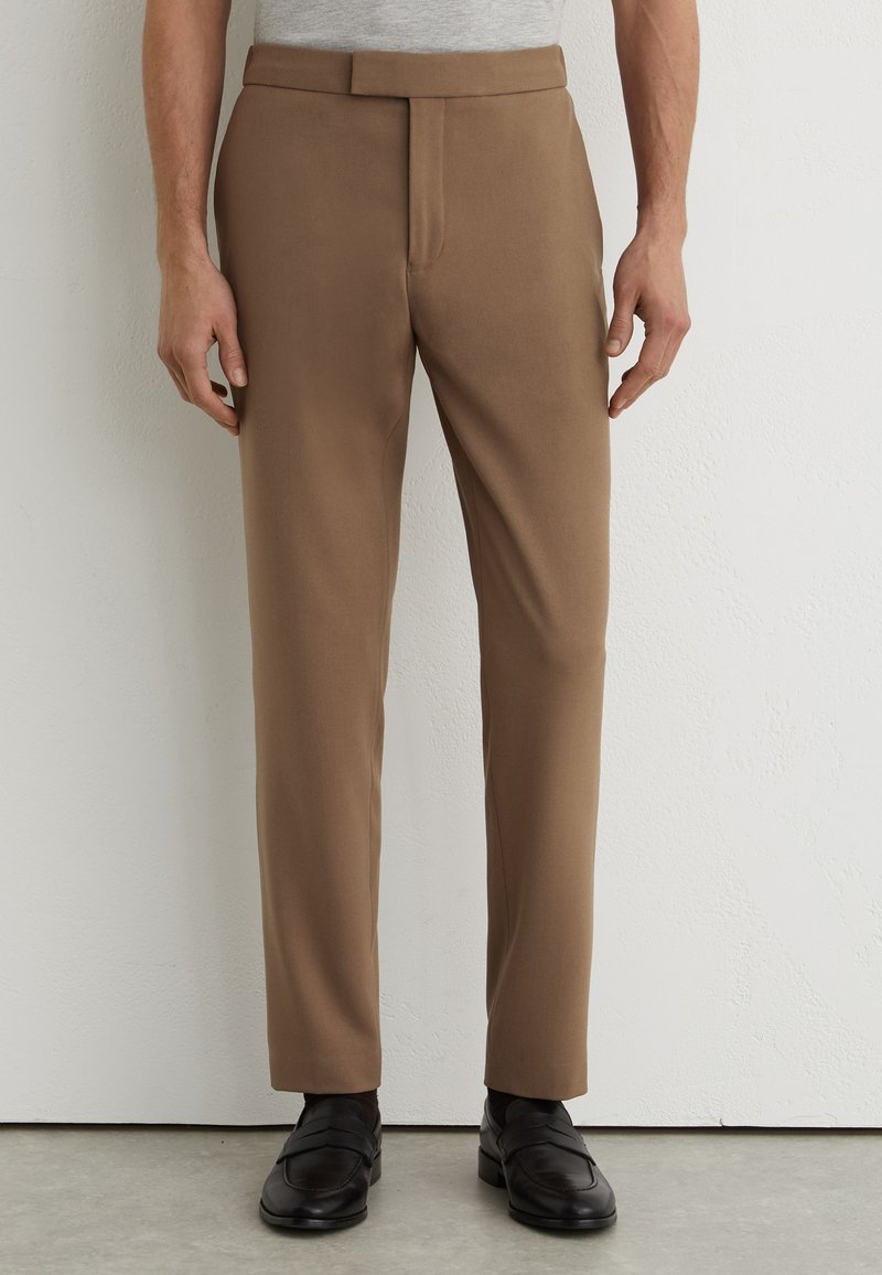 Homme portant un pantalon marron slim fit à devant plat et des mocassins en cuir noirs, debout contre un mur blanc.