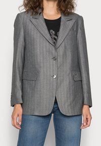 Grijze blazer met visgraatpatroon en pieklapels, twee voorzakken, enkele knoopsluiting en gestructureerde stof. Draagt over een zwart shirt en blauwe jeans.