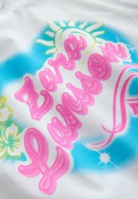 Texte en style graffiti « Zayla Lindsay » en rose avec des motifs soleil et fleurs en bleu, jaune et vert sur tissu blanc.
