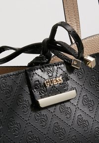 Sac à main Guess noir texturé avec motif de logo embossé, quincaillerie dorée et doublure intérieure beige, montrant des anses nouées.