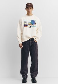 Crèmekleurige sweatshirt met een kleurrijke print, gecombineerd met zwarte spijkerbroeken en zwarte schoenen. Model draagt een bruine pet. Casual pasvorm.