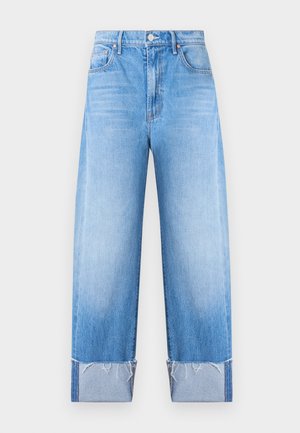 Naine, kellel on pikad mustad juuksed, musta varrukateta topi, kõrge vöökohaga ääristatud siniste teksade ja mustade kontsaga sandaalidega, seisab tavalise seina ees.