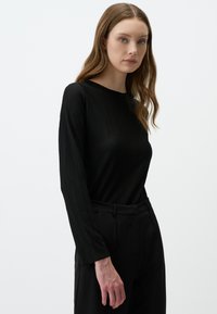 Schwarzes Langarmshirt mit geribbelter Textur. Figurbetontes Design, runder Halsausschnitt. Kombiniert mit hoch geschnittenen schwarzen Hosen, beide in einfarbigen Farben.