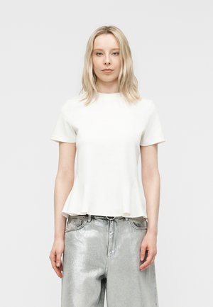 Blond kvinne iført en tettsittende hvit topp med korte ermer og løse metalliske sølvbukser, stående mot en ensfarget grå bakgrunn.