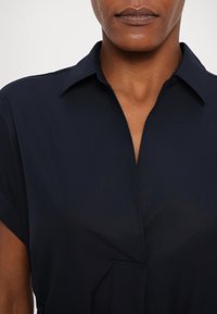 Chemise col marin de couleur bleu marine à manches courtes, avec un décolleté en V peu profond et une texture subtile. Lignes épurées sans motifs ni accents supplémentaires.