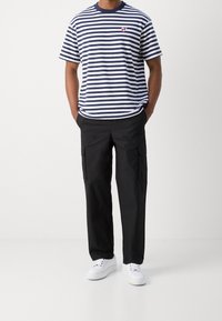 Marineblå og hvid stribet t-shirt med rund hals og lille rød logo, parret med sorte cargo bukser og hvide sneakers.