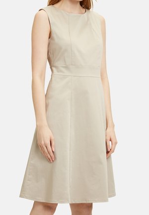 Béžové šaty bez rukávů délky ke kolenům s přiléhavým pasem a kulatým výstřihem, které má na sobě stojící osoba na jednobarevném pozadí.