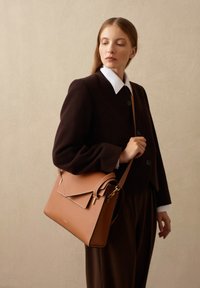 Borsa a mano in pelle marrone con un design strutturato, dotata di chiusura a patta e dettagli in oro. Texture liscia, abbinata a un outfit scuro.