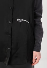 Melna pogu krekls, izgatavots no gludas auduma ar vieglu spīdumu. Uz krūšu ir baltā izšuvumā teksts "KARL LAGERFELD JEANS".