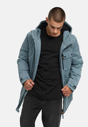 Giacca isolante blu con cappuccio, chiusura a zip e a bottoni, tasche frontali e tessuto testurizzato. Indossata sopra una maglietta nera.