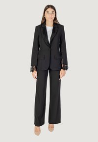 Completo elegante nero con giacca a un bottone con dettagli in pizzo sulle maniche, abbinato a pantaloni a gamba larga e tacchi nudi.