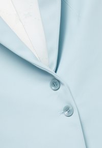 Blazer bleu clair en tissu lisse, avec deux boutons ronds et un col cranté. L'intérieur présente une doublure à motif.