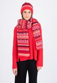 Pull en tricot rouge avec un motif texturé, bonnet rouge assorti avec des rayures multicolores, et une écharpe à motifs avec des designs géométriques. Pantalons noirs.
