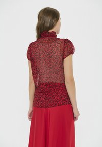 Top rojo con mangas cortas abullonadas; tejido transparente con estampado de leopardo negro; cuello alto con volantes; usado con una falda roja sólida.