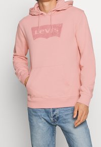 Sweat à capuche rose avec une poche frontale et un logo Levi's texturé dans une couleur contrastante. Tissu doux, coupe décontractée et capuche ajustable avec cordon.