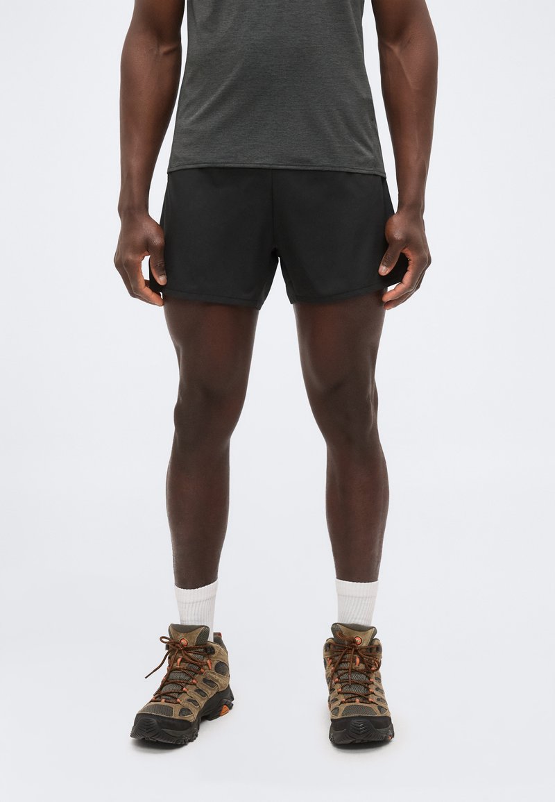 Άνδρας φοράει μαύρα αθλητικά σορτς, γκρι μπλουζάκι, λευκές κάλτσες και καφέ ορειβατικά παπούτσια, στέκεται μπροστά σε απλό φόντο.