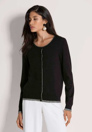 Vrouw met lange, donker haar draagt een zwarte, dichtgeknoopte cardigan met zilveren bies en grote pareloorbellen, gecombineerd met een witte broek.