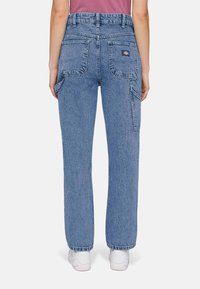 Vista traseira de uma pessoa a usar calças de trabalho de ganga azuis de cintura alta, com grandes bolsos de chapa, um suporte lateral para ferramentas e sapatilhas brancas.