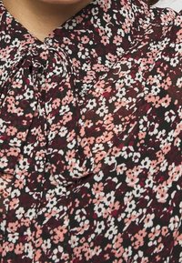 Mujer vistiendo una blusa negra con pequeño estampado floral en rosa, blanco y burdeos, con un lazo atado en el cuello.