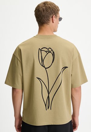 Mann trägt beige übergroßes T-Shirt mit großer schwarzer Linienzeichnung einer Tulpe auf dem Rücken, von hinten gezeigt.