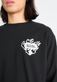 Czarna koszulka z długim rękawem z białym wzorem graficznym przedstawiającym płomienie oraz napisem "MANASTASH" po lewej stronie klatki piersiowej. Materiał: bawełna.