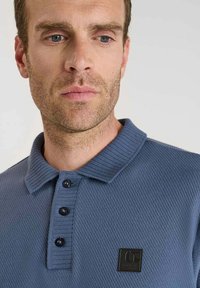 Polo bleu à côtes avec un col à trois boutons, un tissu texturé et un petit patch logo noir sur le côté gauche de la poitrine.