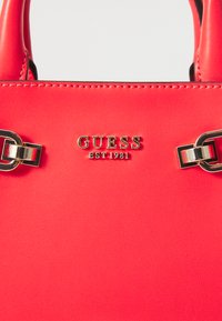 Rød syntetisk taske med et guld "GUESS EST 1981" logo og en metallisk kædedetalje på håndtaget. Glat tekstur med syede detaljer.