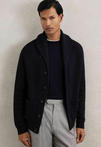 Hombre vestido con un cárdigan negro abotonado sobre una camiseta negra y pantalones de tiro alto gris claro, de pie contra una pared lisa y clara.