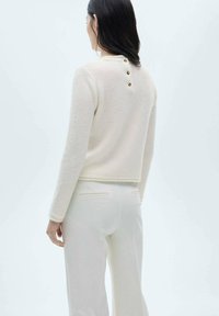 Maglione crema, testurizzato, con design corto, dotato di tre bottoni dorati sul retro, abbinato a pantaloni bianchi, sartoriali.