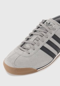 Pelēkas zamšādas tenisbikses ar trim melniem svītrām, strukturētu brūnu gumijas zoli un plakanām pelēkām auklām. Dizains ir pievilcīgs un sportisks.