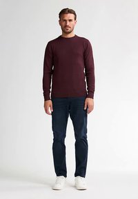 Weinroter Langarm-Pullover mit Rundhalsausschnitt, kombiniert mit dunkelblauen Jeans und weißen Sneakern. Einfarbig, körperbetontes Design und minimale Texturdetails.