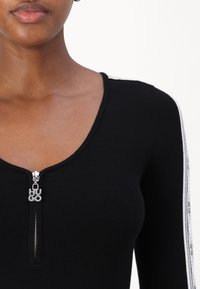 Schwarzes, geripptes Oberteil mit V-Ausschnitt und Reißverschluss, versehen mit einem silbernen Logo-Zipper. Weiße Streifen entlang der Ärmel verleihen dem Design das gewisse Etwas.