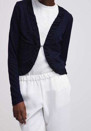 Personne portant un blazer bleu marine foncé avec des bords froncés sur un haut blanc texturé et un pantalon blanc ample sur un fond uni.
