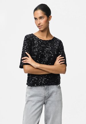 Woman with dark hair in a black sequined short-sleeve top and light gray jeans, standing with arms crossed against a plain white background.
