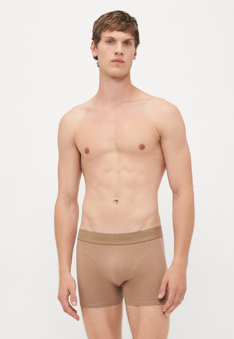 Jonge man die beige boxershorts met een merkband draagt, staand tegen een effen lichte achtergrond, met het gezicht naar voren.
