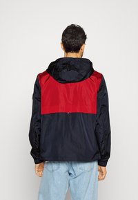 Giacca resistente all'acqua con sezione superiore rossa e parte inferiore blu navy. Include un cappuccio e polsini elastici per una vestibilità sicura.