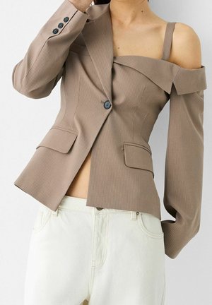 Asymmetrische beige pinstripe blazer met één blote schouder en bijpassende band, gedragen met witte broek met hoge taille.