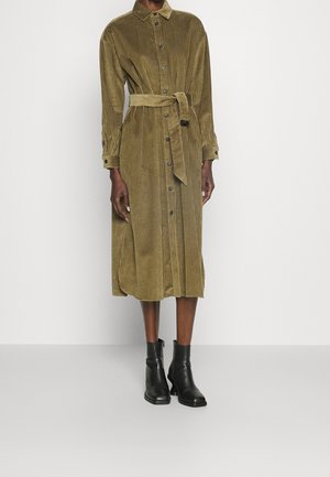 Osoba o ciemnej karnacji ubrana w oliwkowozieloną sukienkę koszulową z kożuchem do kolan z paskiem i czarne botki, stojąca na tle jednolitej ściany.