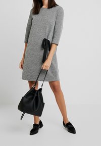 Robe tricotée grise à manches trois-quarts, avec un détail de ceinture noire à nouer à la taille, accompagnée d'un sac seau noir texturé et de chaussures noires.